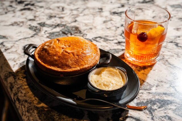 Glazed skillet cake served with whipped butter alongside an old fashioned cocktail garnished with an orange twist and cherry on a marble tabletop.