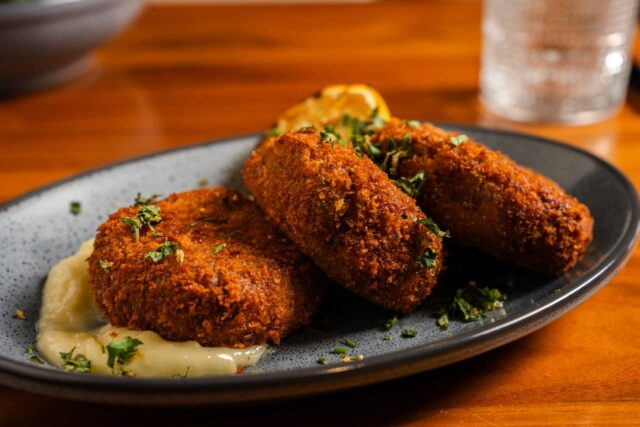 Three golden-brown crab cakes served with a creamy dipping sauce and lemon wedge, garnished with fresh herbs on a dark plate.