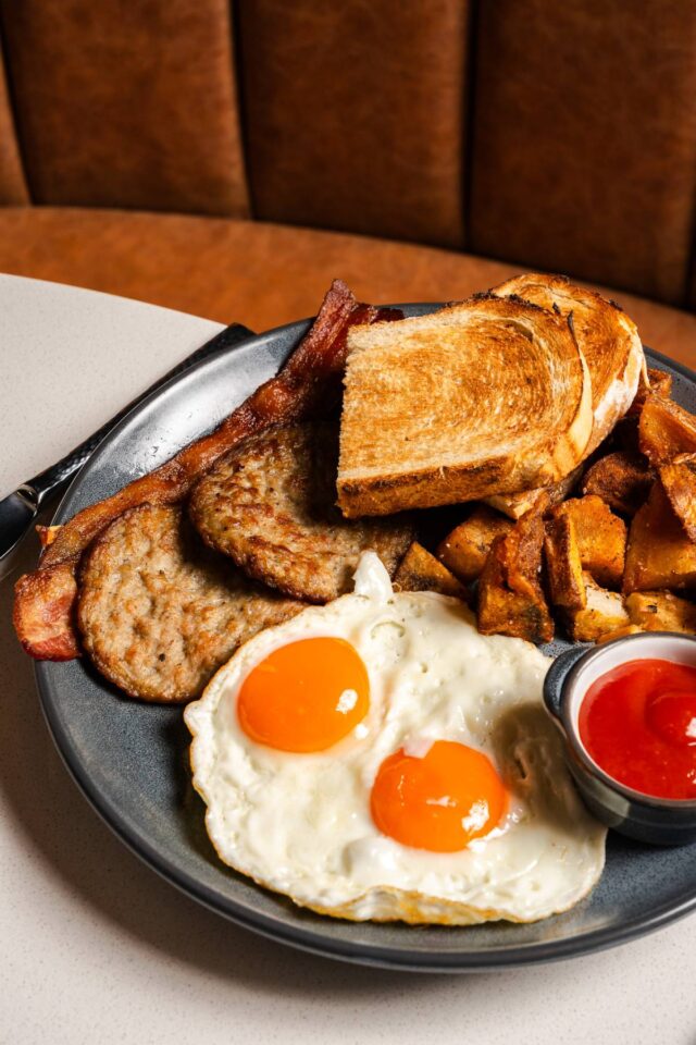 Traditional breakfast plate with two sunny-side-up eggs, bacon, breakfast sausage, roasted potatoes, toasted bread, and a side of ketchup served on a dark ceramic plate.