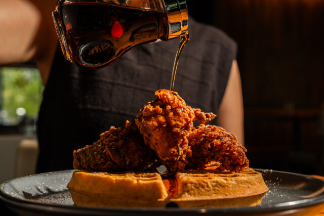 Crispy fried chicken served over a golden waffle as maple syrup is poured from above, creating a warm and indulgent chicken and waffles presentation.