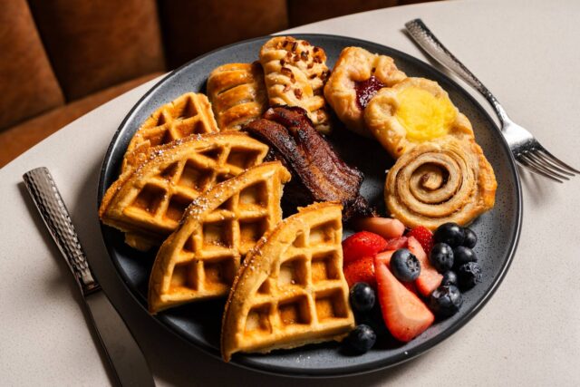 Breakfast plate featuring golden waffles, crispy bacon, assorted pastries, fresh strawberries and blueberries, served on a dark ceramic plate with knife and fork.