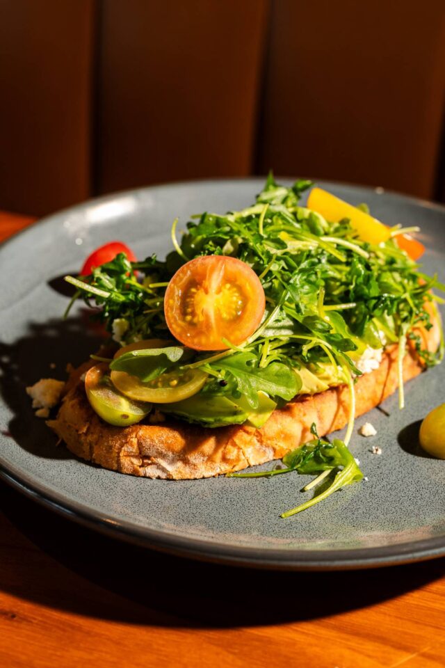 Avocado toast topped with fresh arugula, cherry tomatoes, and crumbled cheese, served open-faced on toasted artisan bread.