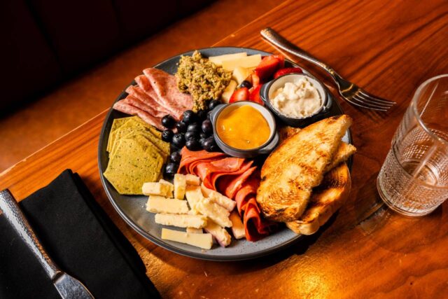 Charcuterie board featuring assorted cheeses, cured meats, crackers, toasted bread, fresh berries, and spreads, served on a wooden restaurant table.