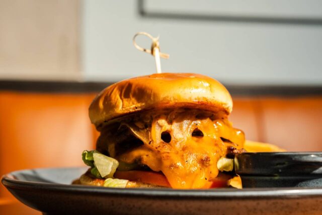 Close-up of a cheeseburger topped with melted cheddar cheese, lettuce, tomato, and pickles on a toasted bun, served with fries and dipping sauce.