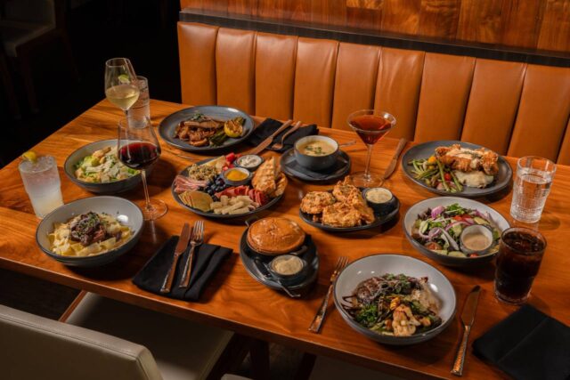 Wide view of a restaurant table set with a variety of dishes including pasta, grilled steak, fried chicken, salads, charcuterie, soup, dessert, and cocktails, surrounded by wine and soft ambient dining room lighting.