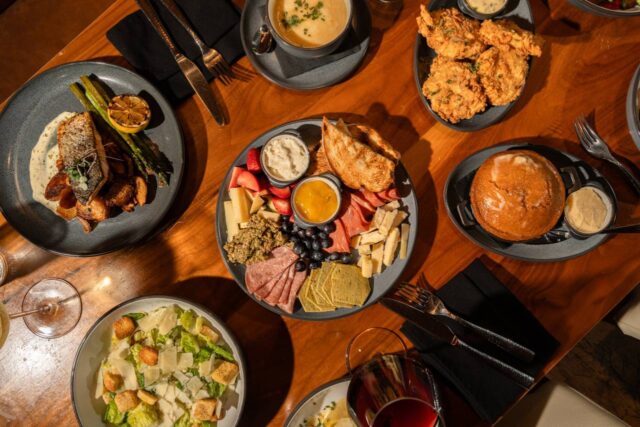 Overhead view of a restaurant table featuring a charcuterie board with cheeses, cured meats, fruit and crostini, crispy fried chicken, creamy soup, grilled fish with asparagus, Caesar salad, glazed skillet cake with whipped butter, and glasses of wine in warm natural light.