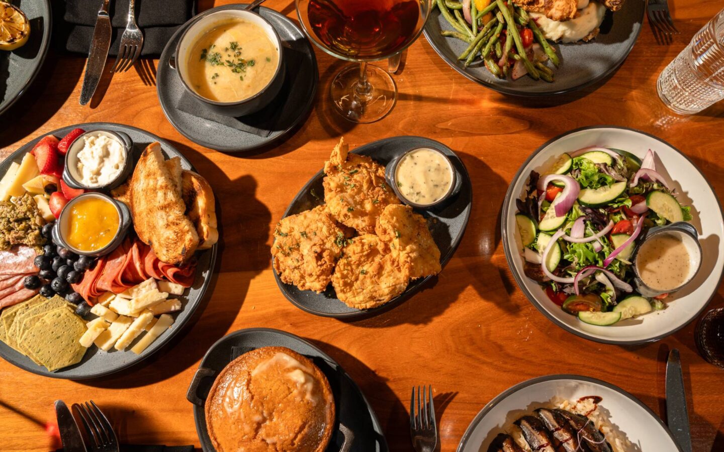 Overhead view of a restaurant table featuring a charcuterie board with cheeses, cured meats, fruit and toast, crispy fried chicken with dipping sauce, a fresh garden salad, creamy soup, a plated entrée with vegetables, a glazed pastry, and a glass of red wine in warm natural light.