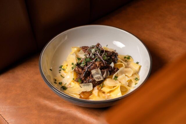 Short Rib Pappardelle featuring braised beef, wide ribbon pasta, shaved parmesan, and microgreens, served in a white bowl on a leather banquette seat.