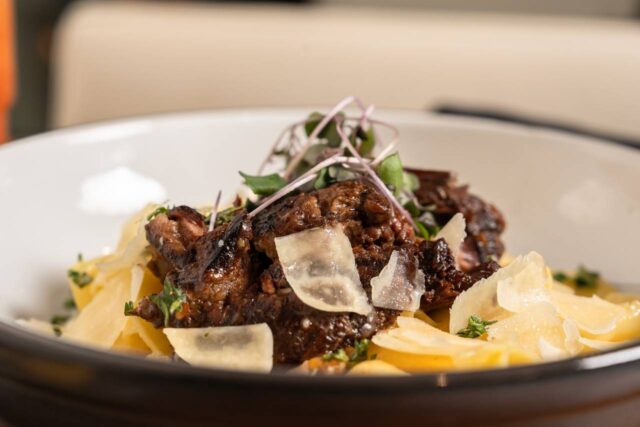 Braised beef pasta topped with shaved parmesan and microgreens, served in a white bowl with wide ribbon noodles and rich savory sauce.