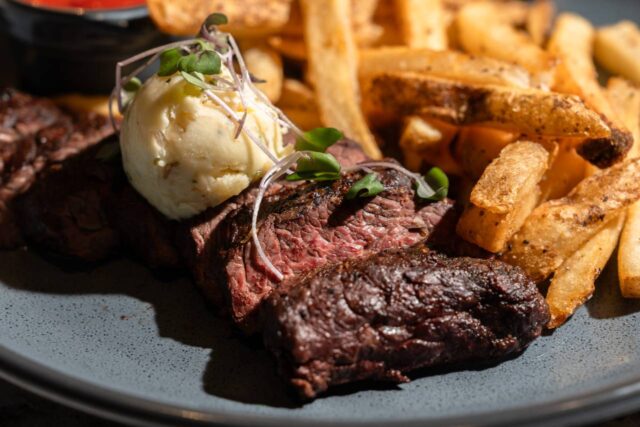 Grilled steak sliced and served with seasoned fries, topped with a scoop of herb butter and microgreens on a dark plate.