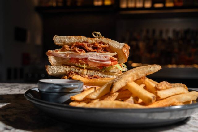 Club sandwich stacked with sliced turkey, bacon, lettuce, tomato, and cheese on toasted bread, served with seasoned fries and a side of dipping sauce.