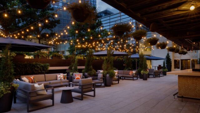 Outdoor restaurant patio with cushioned lounge seating, lush greenery, hanging planters, and warm string lights glowing at dusk.