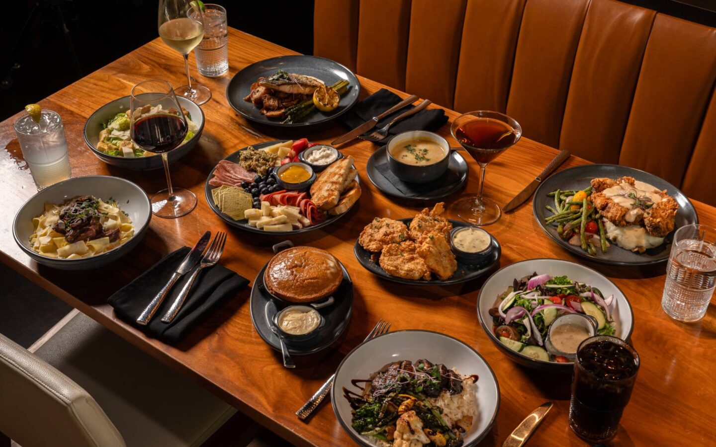 Assorted restaurant dishes, including pasta, salad, soup, fried chicken, grilled meat, and a charcuterie board, served with wine and cocktails on a wooden table.