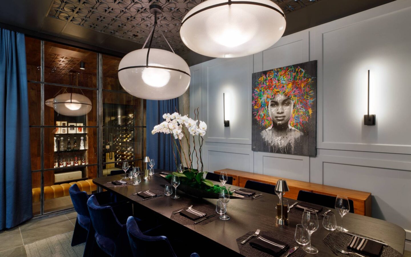 Private dining room with a set table, blue velvet chairs, pendant lights, orchids, wine storage, and colourful wall art.