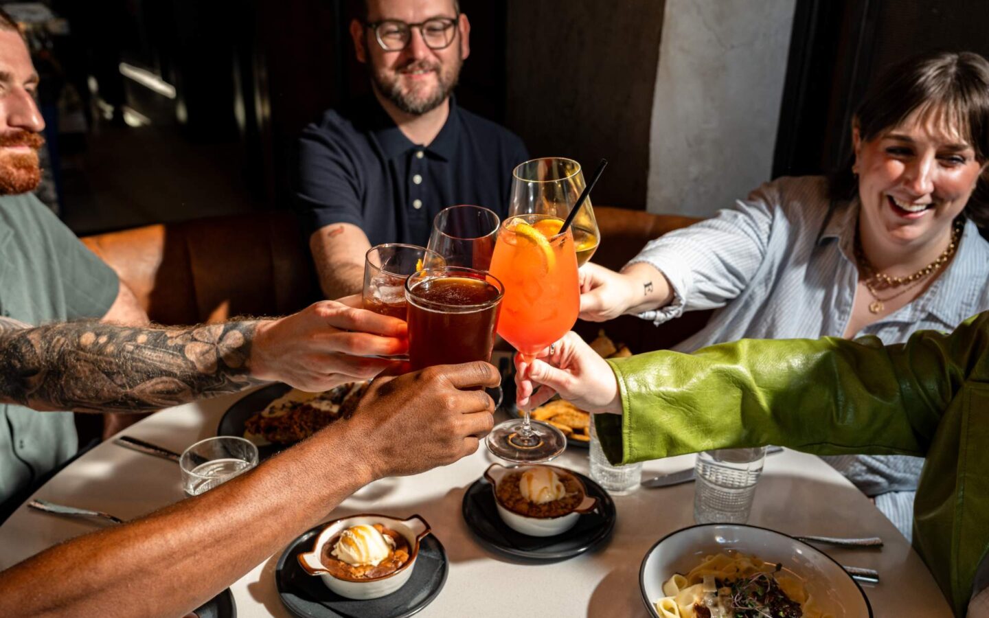 Friends raising cocktails in a toast over a restaurant table with food and desserts.