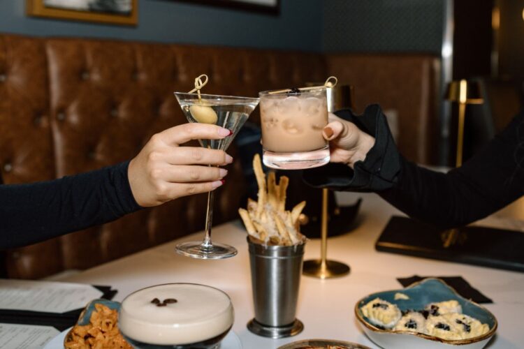 Two people toasting cocktails over a table with food.