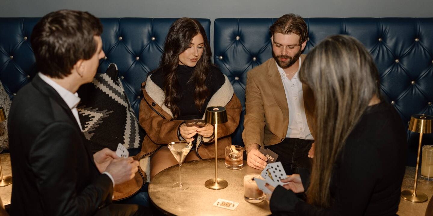 Four people playing cards around a table with cocktails in a stylish lounge featuring blue tufted booth seating and warm ambient lighting.