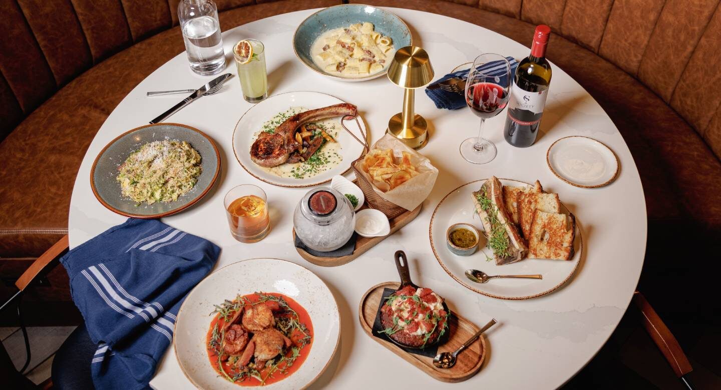 Overhead view of a restaurant table set with a variety of dishes, including a grilled pork chop, pasta, sandwiches, appetizers, chips, cocktails, red wine, and water in a cozy booth setting.