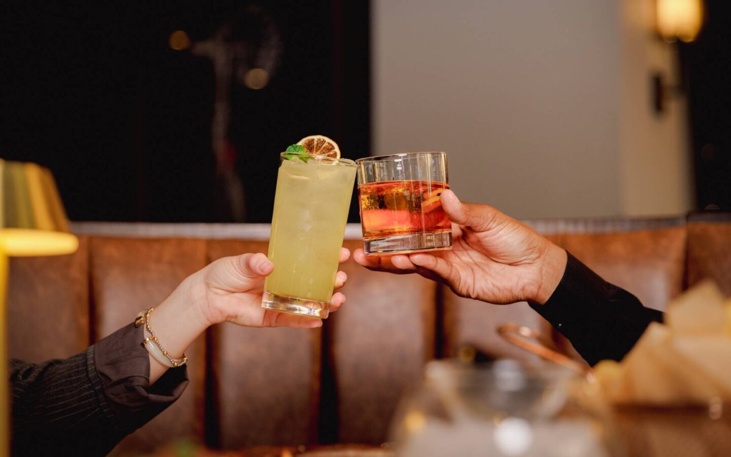 Two people toasting with a yellow cocktail and an amber mixed drink across a restaurant table in a warmly lit dining room.
