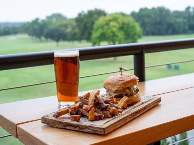 burger and fries on a wooden board next to a glass of beer overlooking the golf course
