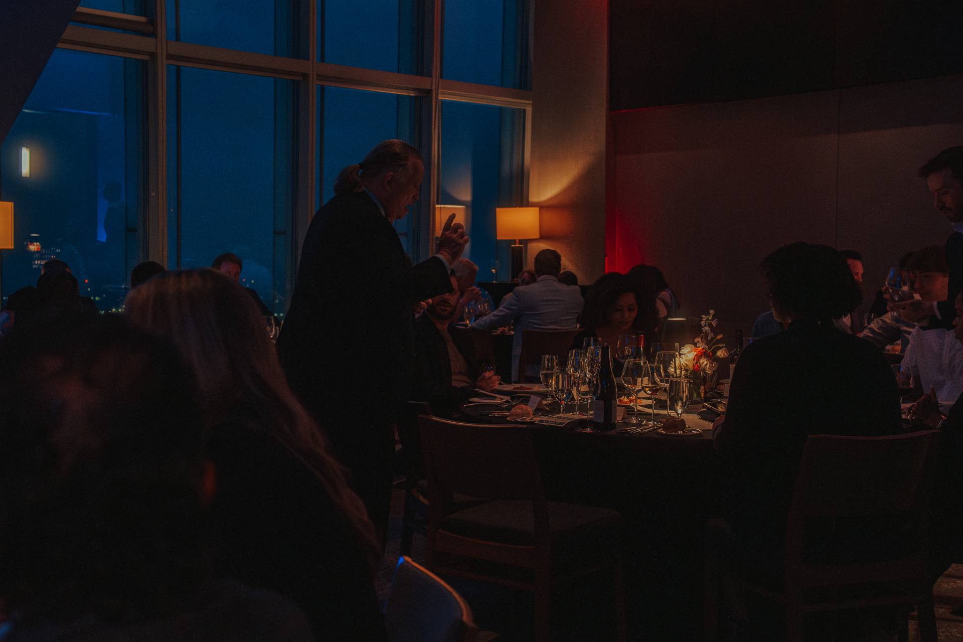 Guests dining in a dimly lit restaurant while a server attends to a table beside floor-to-ceiling windows at night.