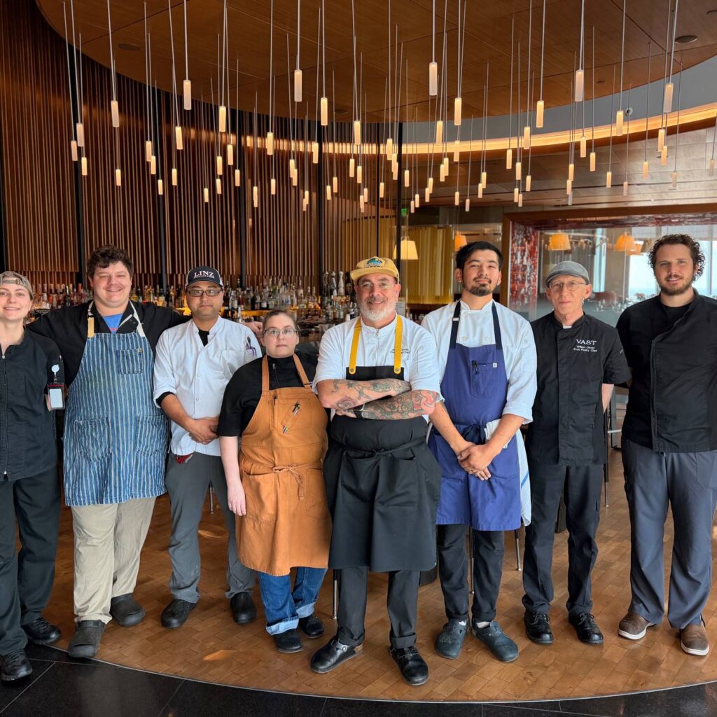 Group of chefs posing together inside a modern restaurant dining room with a bar and hanging lights.