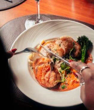 A close-up shot of a diner using a fork and knife to slice into a chicken roulade served with vegetables and a vibrant sauce in a white bowl.