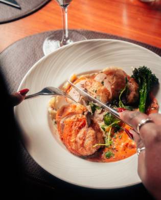 A close-up shot of a diner using a fork and knife to slice into a chicken roulade served with vegetables and a vibrant sauce in a white bowl.