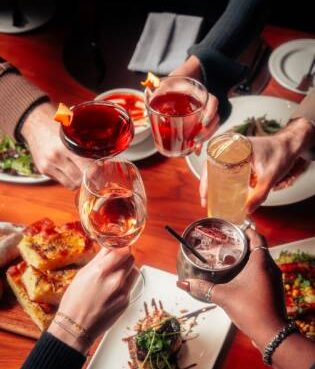 A group of people clinking various colorful cocktails and drinks together in a toast over a wooden restaurant table filled with small plates and appetizers.