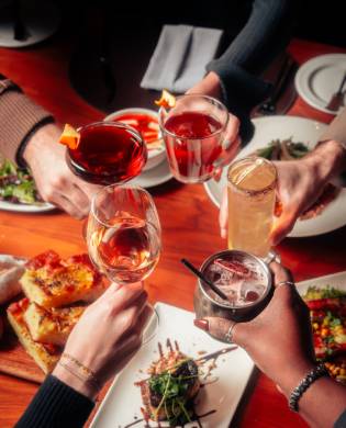 A group of people clinking various colorful cocktails and drinks together in a toast over a wooden restaurant table filled with small plates and appetizers.