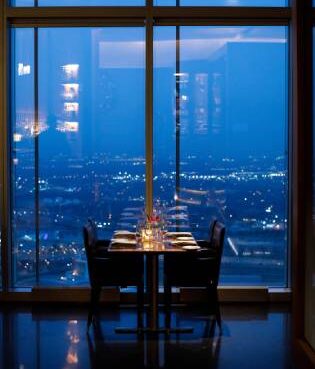 A dining table set for two with glowing candles and wine glasses, positioned next to floor-to-ceiling windows overlooking a panoramic, illuminated city skyline at night.