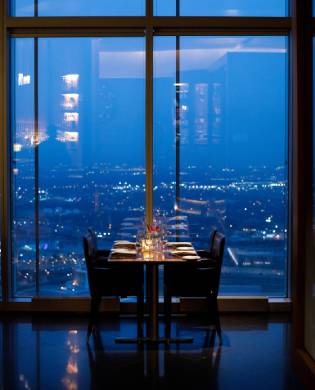A dining table set for two with glowing candles and wine glasses, positioned next to floor-to-ceiling windows overlooking a panoramic, illuminated city skyline at night.