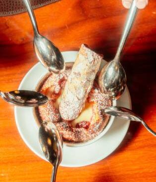 A top-down view of a tart cherry clafoutis dessert topped with powdered sugar and a pastry, surrounded by multiple spoons dipping into the dish on a wooden table.