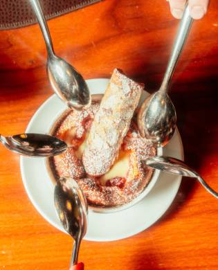 A top-down view of a tart cherry clafoutis dessert topped with powdered sugar and a pastry, surrounded by multiple spoons dipping into the dish on a wooden table.
