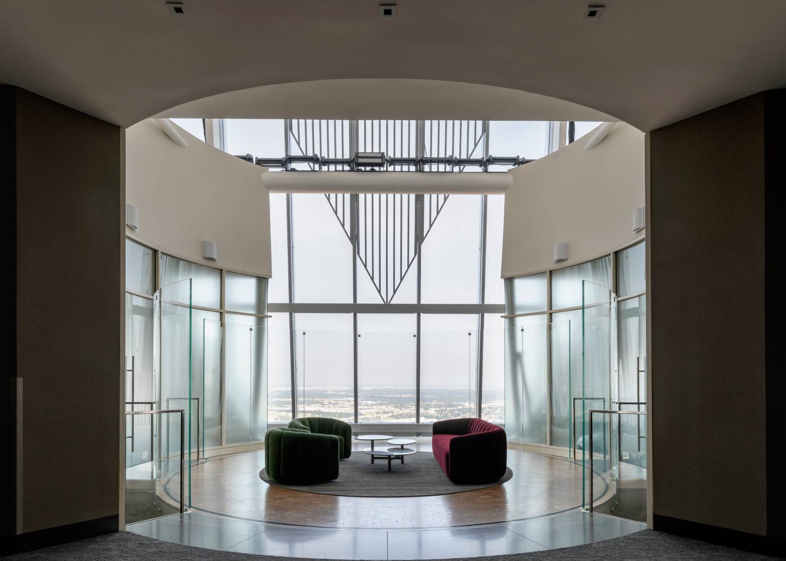 Modern lounge area with curved glass walls, green and burgundy seating, and floor-to-ceiling windows overlooking the city.