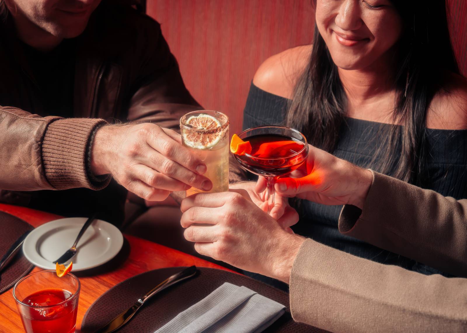 Three people toasting with cocktails over a restaurant table in warm red lighting.