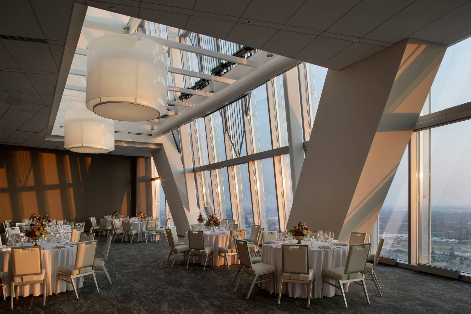 Elegant event space with round dining tables, floral centerpieces and floor-to-ceiling windows overlooking the city at sunset.
