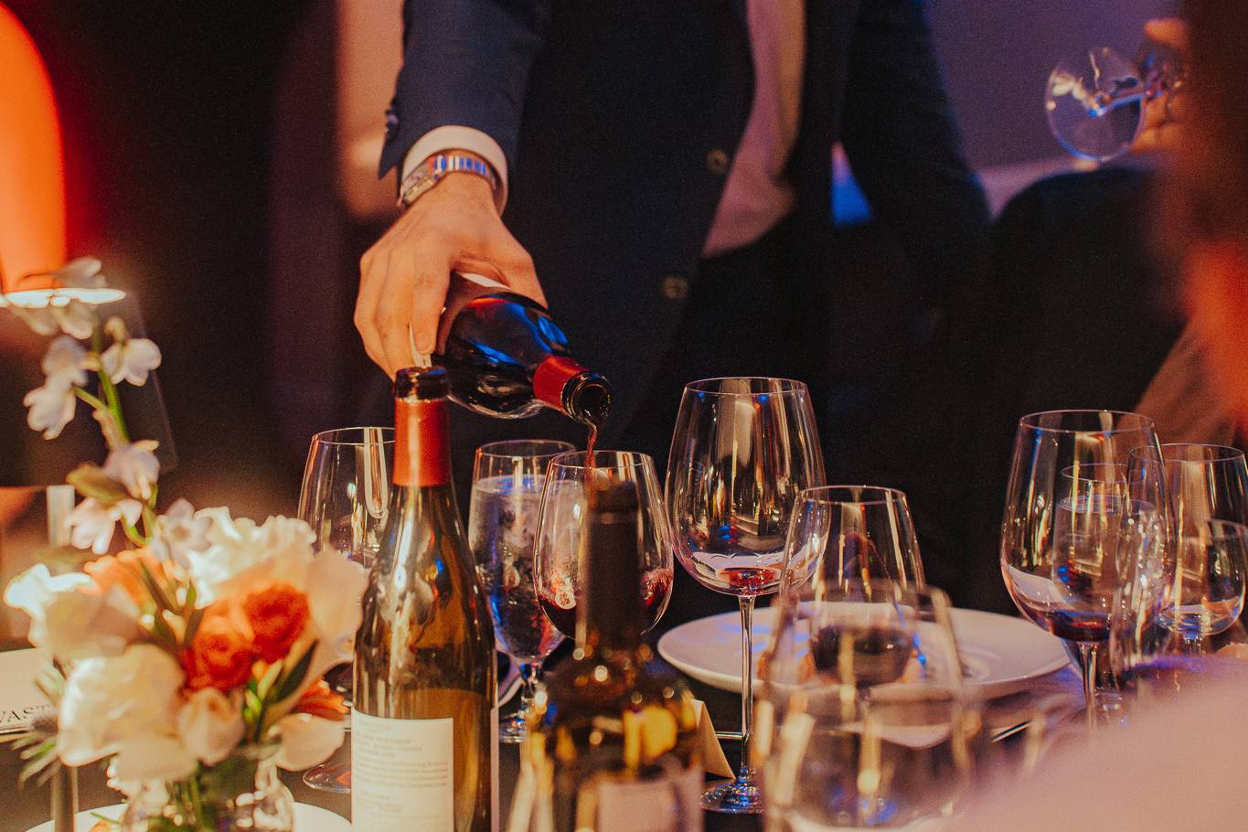Server pouring red wine into a glass at an elegantly set dinner table.