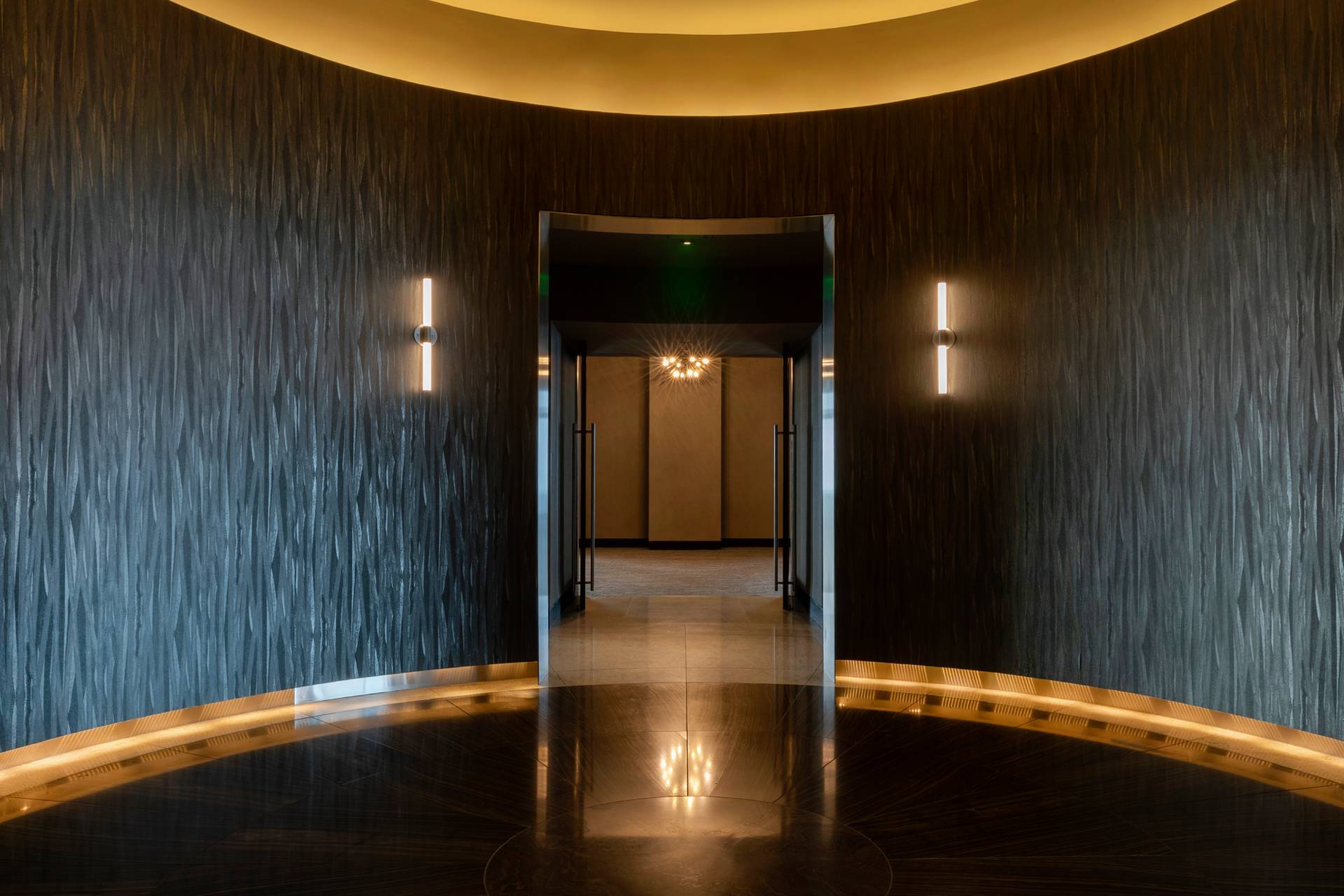Modern hotel corridor with curved dark textured walls, warm recessed ceiling lighting, and polished floors.